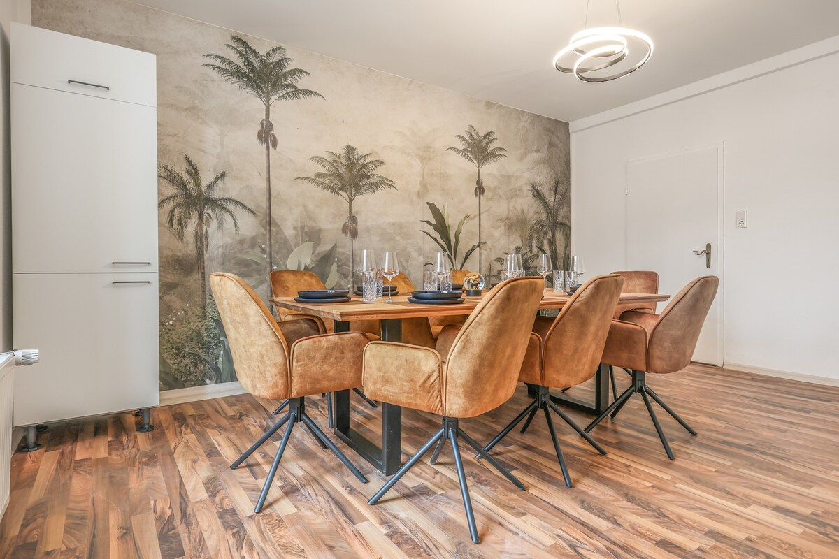 A dining area features a large wooden table surrounded by six plush, brown chairs. The wall is adorned with a mural showcasing palm trees, creating a serene ambiance. Warm wood flooring enhances the overall inviting feel of the space.