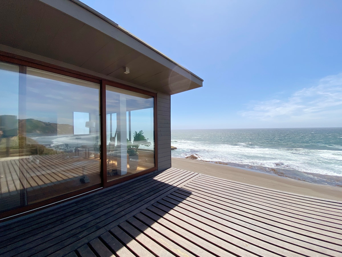 A modern structure is situated on sandy beachfront, with large glass windows offering views of the ocean. A wooden deck extends towards the shoreline, and reflections from the windows reveal coastal scenery and a bright sky.