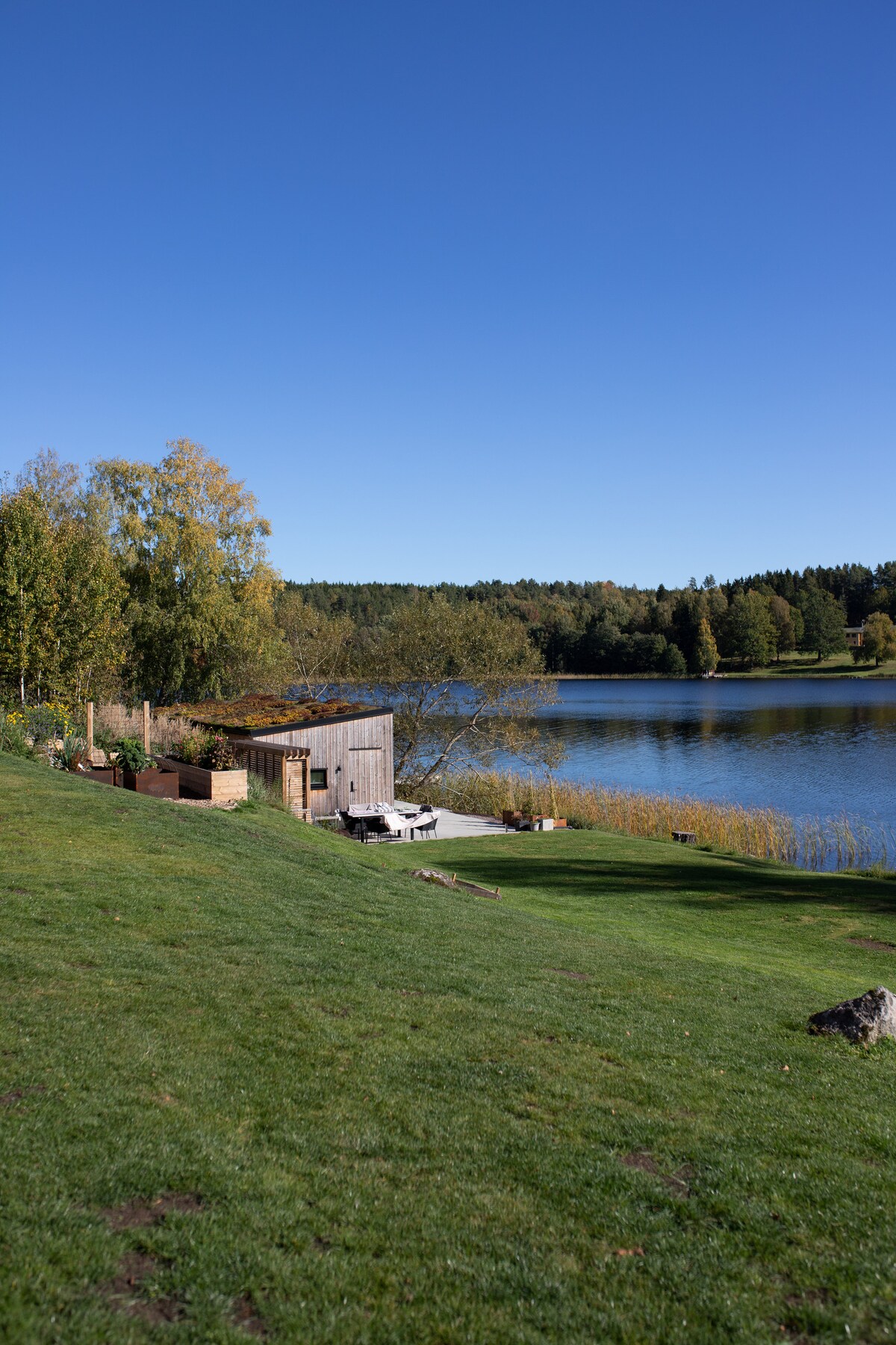 A serene lakeside view showcases a wooden building set against lush greenery and a clear blue sky. The gentle slope leads to the water's edge, with seating arranged on a patio area, inviting relaxation and connection with nature.