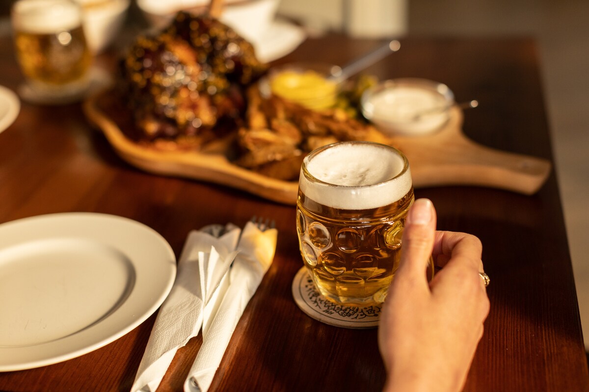A glass of beer is held on a coaster, surrounded by various dishes on a wooden table. In the background, a platter of roasted meat is presented alongside sauces and garnishes. White plates and wrapped cutlery are arranged nearby.