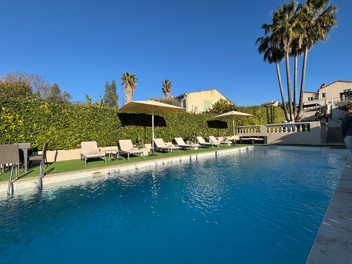 A well-maintained pool area is surrounded by lush greenery, featuring sun loungers and shaded umbrellas. The clear blue water reflects the sunny sky, while nearby palm trees complete the inviting outdoor space, ideal for relaxation and leisure.