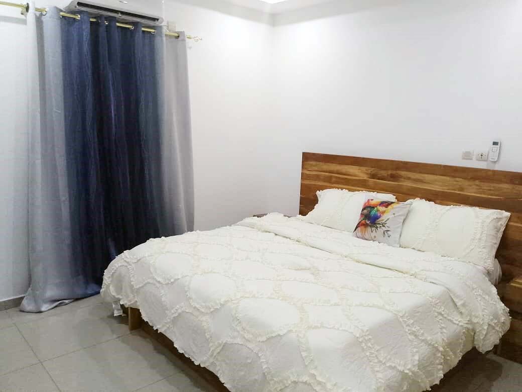 A well-appointed sleeping area features a spacious bed with a textured white coverlet. Soft gray curtains frame a window, allowing natural light to softly illuminate the room. A decorative pillow adds a touch of color to the neutral tones of the space.