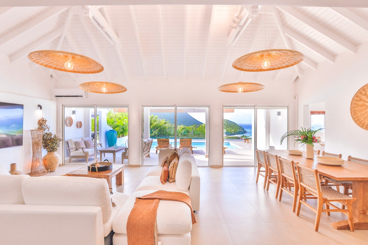 A spacious living area is featured, characterized by high, white ceilings and natural light flowing through large glass doors. Earthy tones from wicker lighting and furniture are complemented by a large dining table and cozy seating, creating an inviting environment near the outdoor pool area.