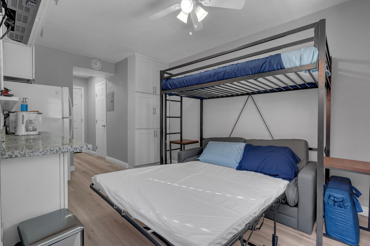 A compact sleeping area is furnished with a twin bunk bed above a full-sized pull-out sofa. Neutral walls and light wood flooring create a calming ambiance. A small kitchenette is visible in the background, featuring a granite countertop and essential appliances.