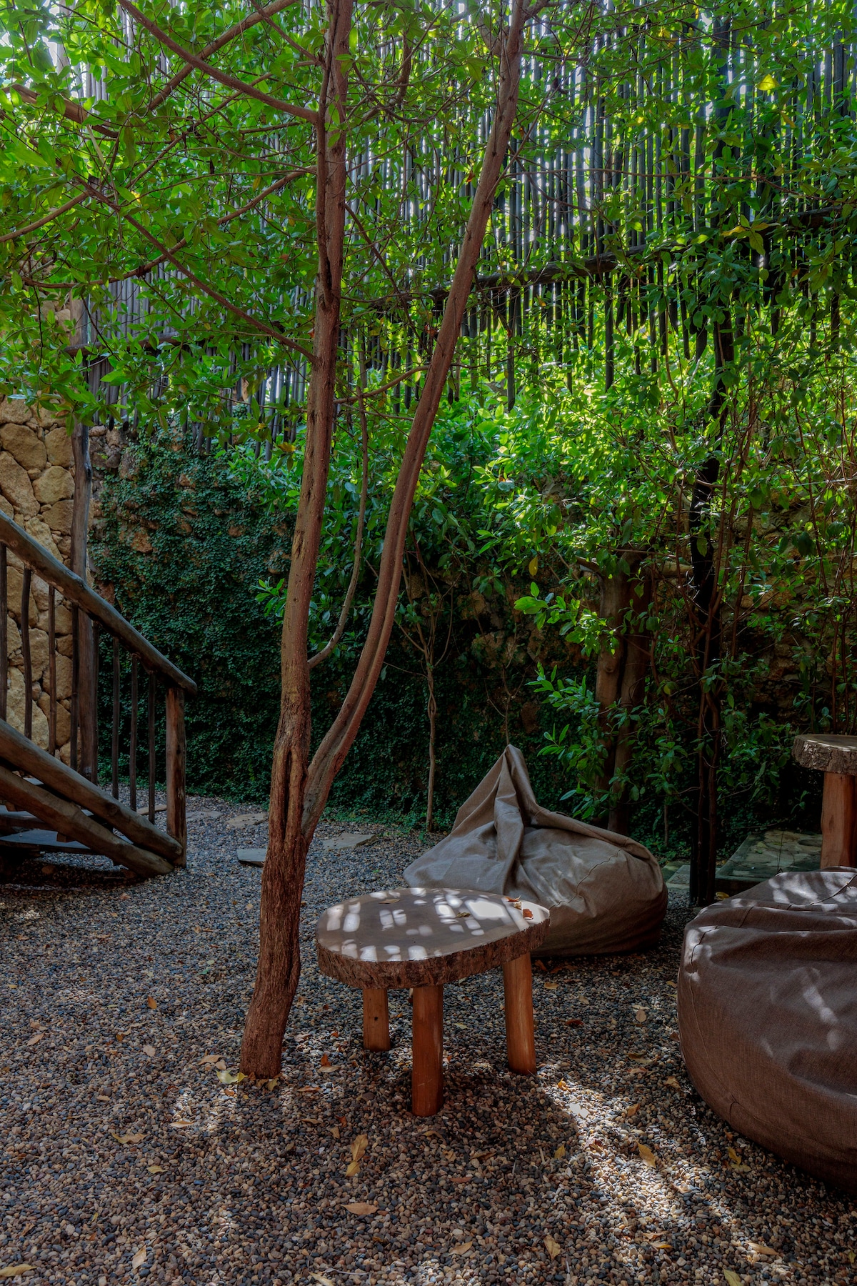 A serene outdoor space is surrounded by lush greenery and filtered sunlight. A small wooden table and comfortable bean bags are positioned on a gravel surface, inviting relaxation. A slender tree provides natural shade, enhancing the tranquil atmosphere.