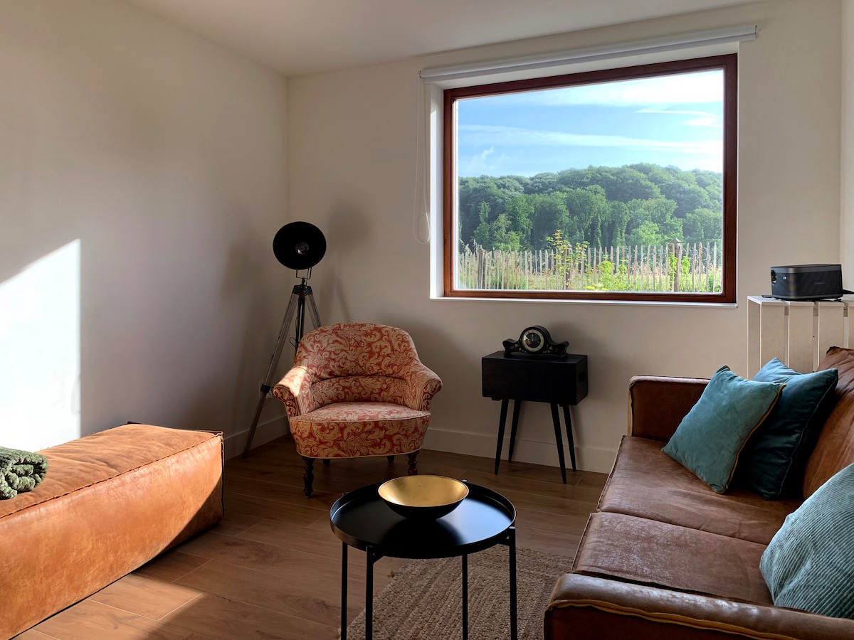 A sunlit living area features a brown leather sofa and an armchair, both positioned near a large window. The window offers a view of the lush greenery outside. A black side table and vintage decor items complement the space.