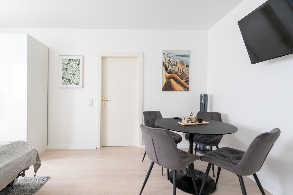 A bright and spacious living area features a round black dining table surrounded by four gray upholstered chairs. A large wall-mounted television is positioned above, and a wall art piece adds visual interest. The door leads to another room, enhancing the open layout.