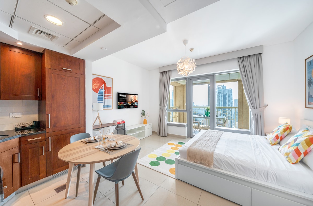 A sleek studio features an open layout with a plush king-size bed, a compact dining table, and a modern kitchenette. Full-length windows allow ample natural light and showcase a view of the balcony. A colorful rug adds a lively touch to the spacious floor.