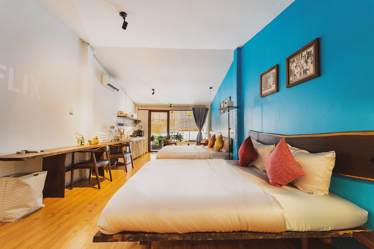 A spacious room features two king-size beds with soft linens and decorative pillows. A long wooden desk spans one side, providing ample workspace. Large windows allow natural light to fill the area, and a balcony with greenery is visible in the background, offering a welcoming outdoor space.
