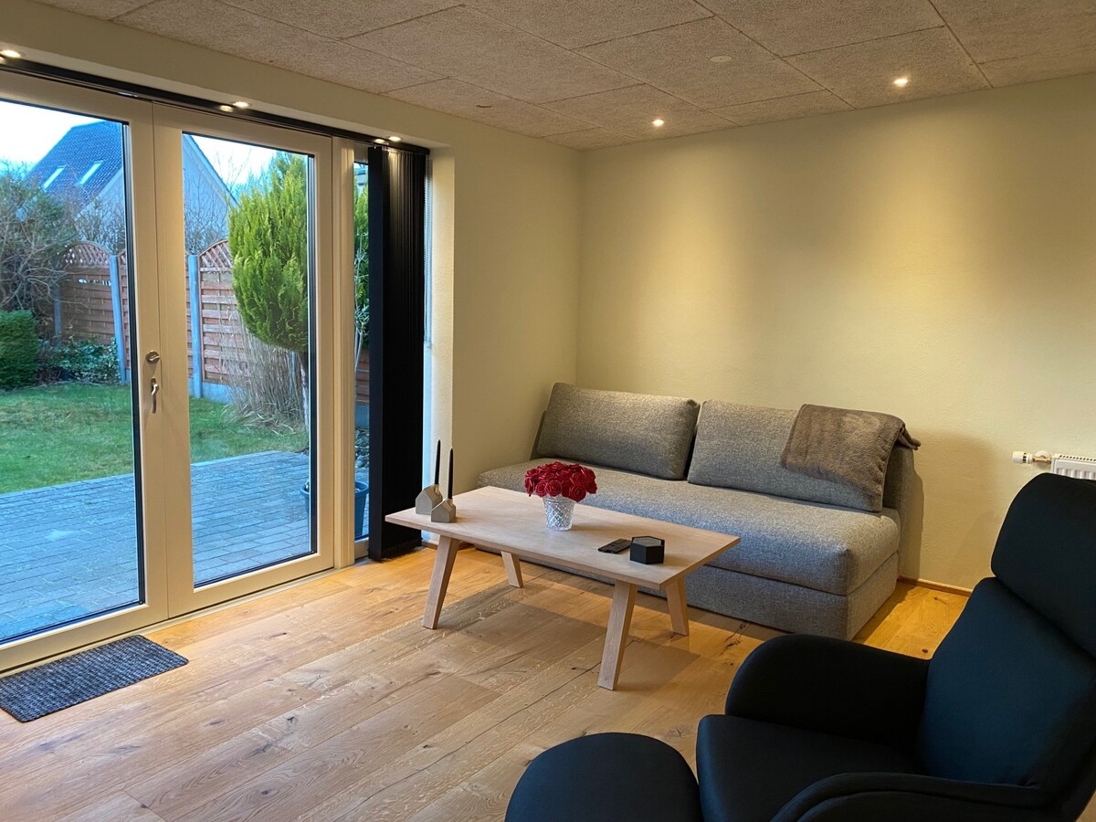 A bright and inviting living area features a spacious gray sofa and a low wooden coffee table adorned with a vase of red flowers. Glass doors open to a private garden space, enhancing natural light and connectivity to the outdoors.