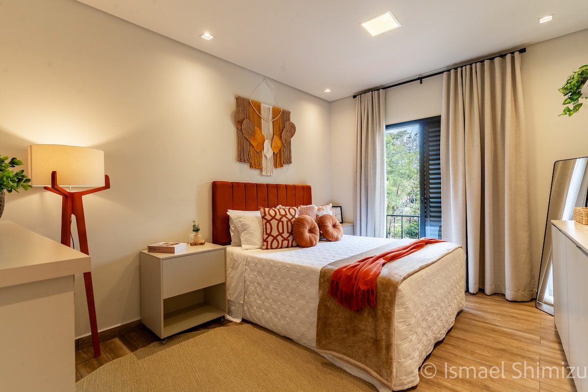 A bright sleeping area features a spacious bed with decorative pillows and a warm throw. Natural light enters through a window with light curtains, and a minimalist bedside table holds a plant. A softly glowing lamp adds a touch of warmth to the neutral-toned decor.