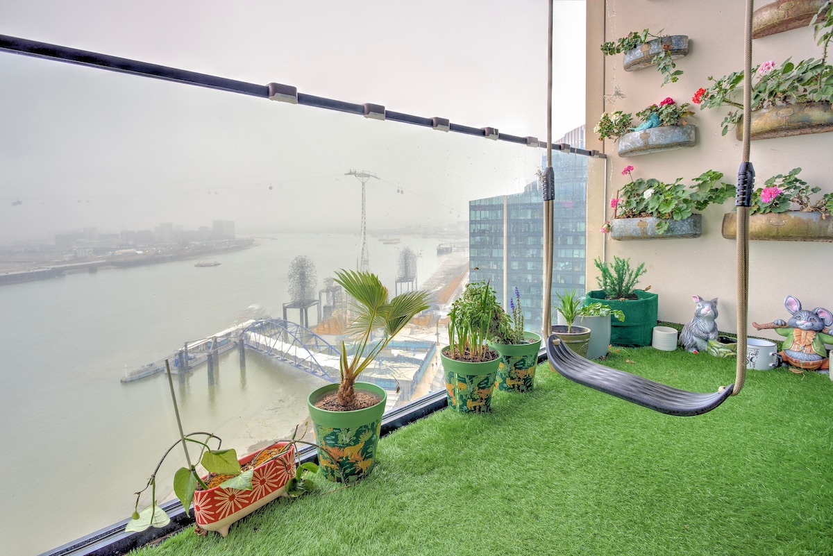 A balcony is embraced by a glass railing, offering views of the river and surroundings. Potted plants in various shapes and sizes decorate the space, while an artificial grass surface adds a touch of greenery. A swing seat hangs, inviting relaxation amid the scenic backdrop.