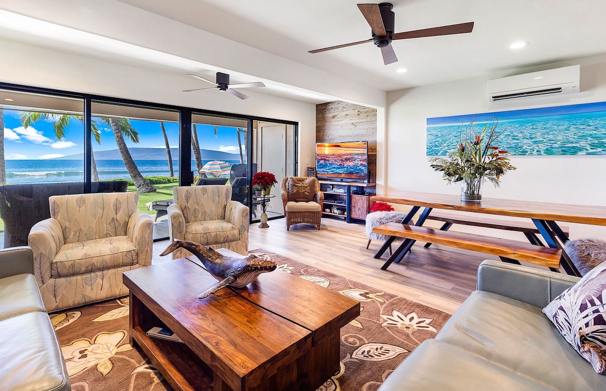 An inviting living space features comfortable seating arranged around a wooden coffee table. Large windows provide uninterrupted ocean views, allowing natural light to illuminate the room. A dining table is visible, showcasing an open layout that seamlessly connects to the outside.