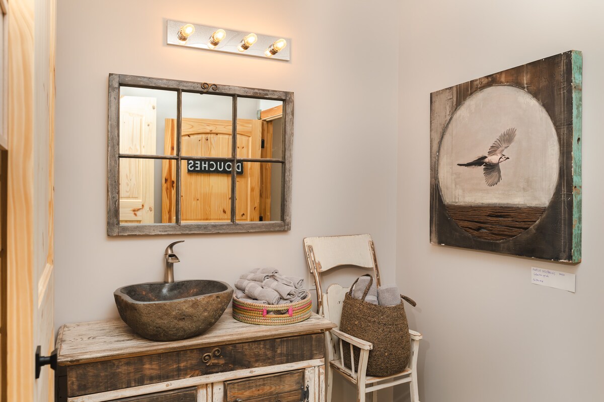 A shared bathroom features a rustic wooden vanity with a circular stone sink. Above, a mirror framed with reclaimed wood reflects soft lighting. A piece of wall art depicting a bird in flight is displayed nearby, enhancing the tranquil ambiance.