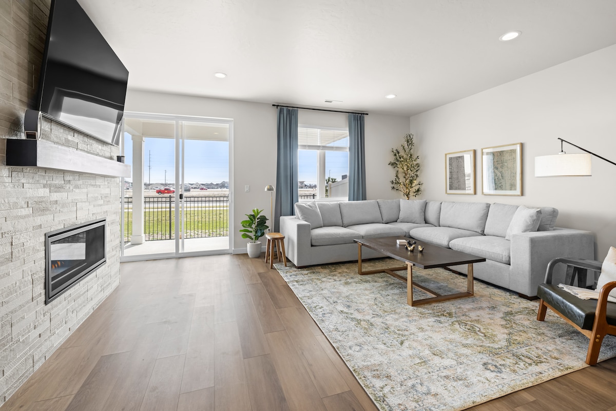 A contemporary living area is highlighted by a comfortable sectional sofa facing a modern fireplace. A large area rug anchors the seating space, while natural light floods in through sliding glass doors leading to the outdoor area. A simple coffee table and potted plant add subtle touches.