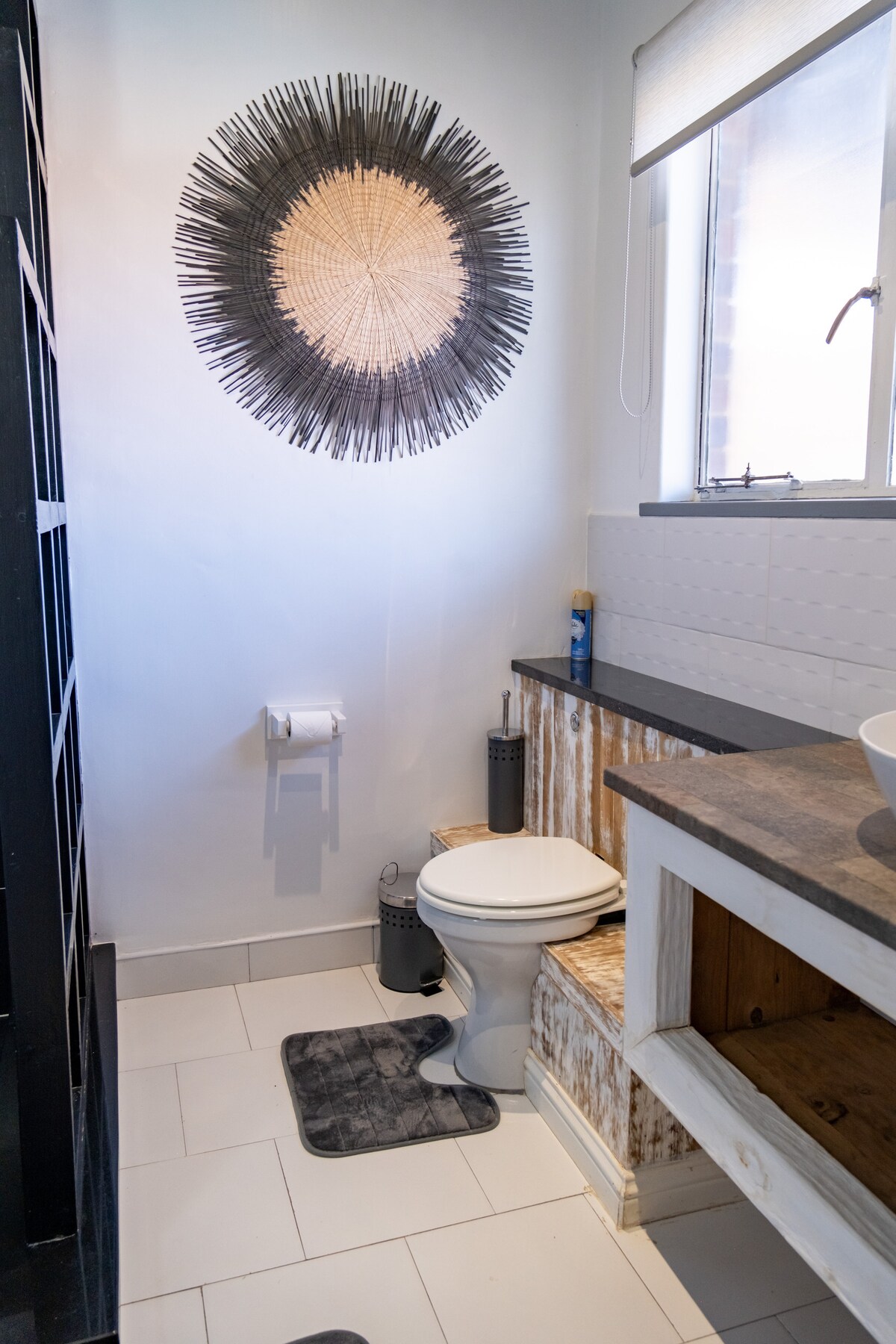 A bathroom is equipped with a modern toilet and a rustic wooden accent on one wall. A circular wall decoration adds texture, while a small dark mat is positioned in front of the toilet. Natural light enters through a nearby window with a light-filtering shade.