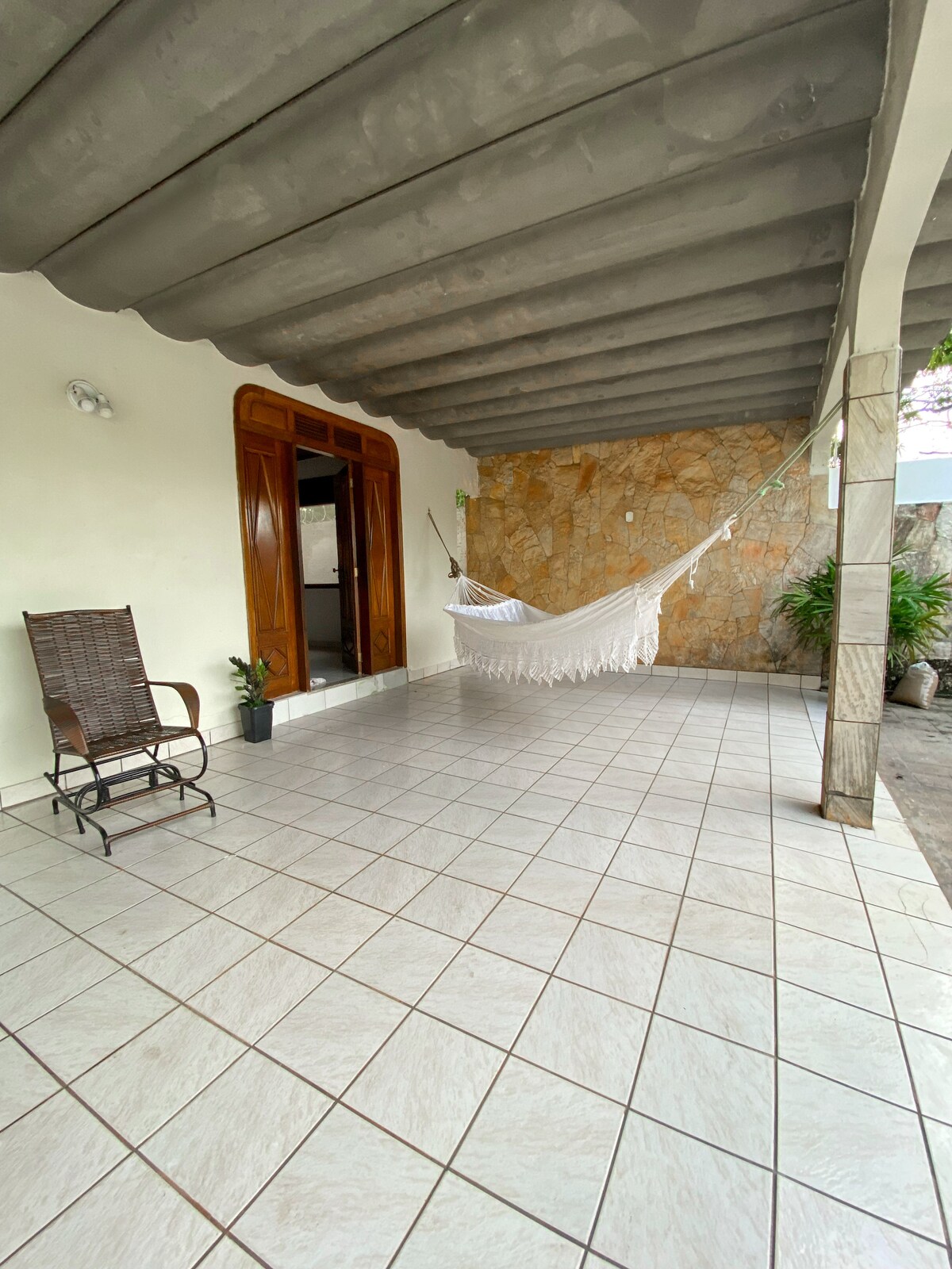 A covered outdoor area is showcased with tiled flooring and natural stone walls. A cozy hammock is positioned against the wall, while a wooden rocking chair is placed nearby. Potted plants add a touch of greenery, enhancing the space's inviting atmosphere.