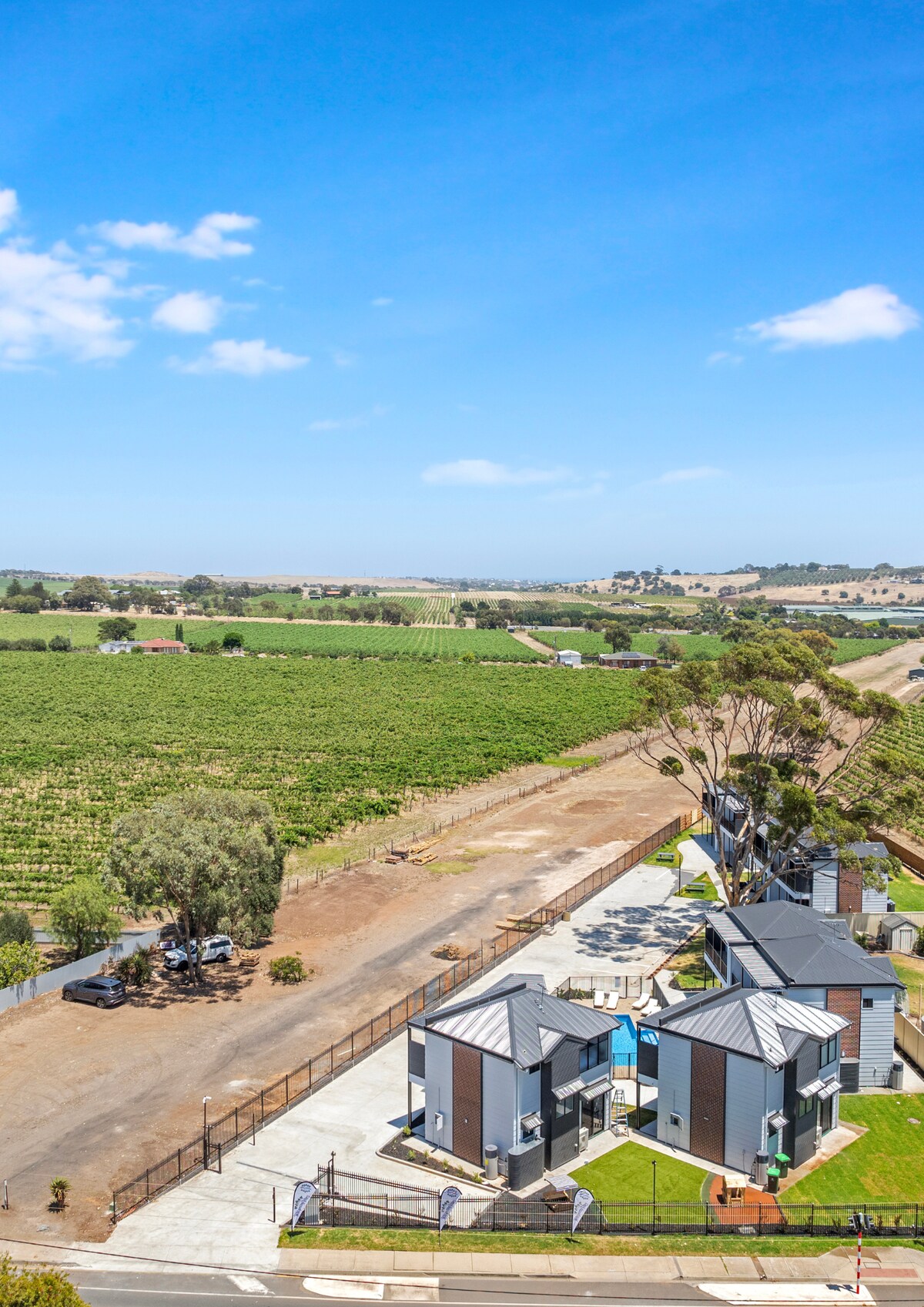 The image presents a view of several modern villas, featuring distinct architectural lines, alongside lush green vineyards that extend into the horizon. A well-maintained, paved driveway provides access, while the surrounding landscape captures the serene beauty of South Australia's wine region.