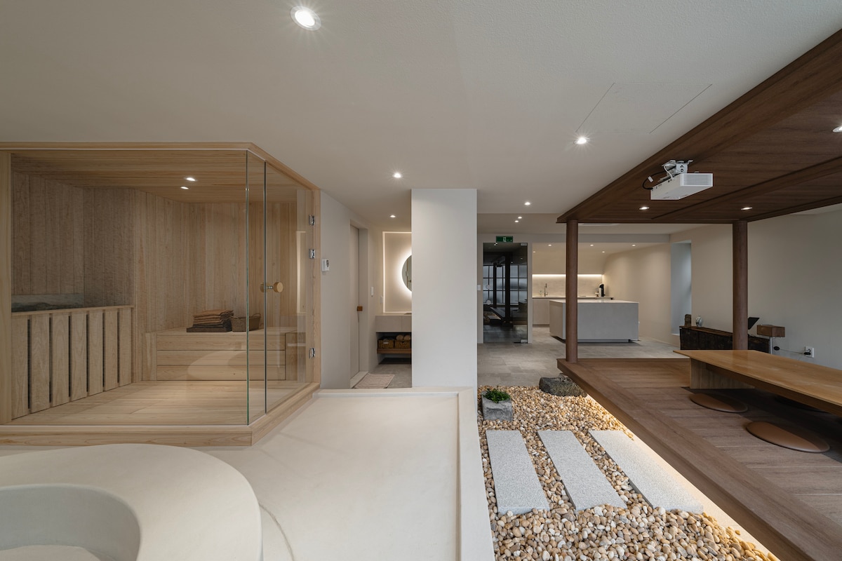 An open and modern interior is highlighted by a glass-enclosed sauna, surrounded by natural wood accents. Neutral tones and soft lighting create a calming environment. A large projection screen is positioned in a spacious area, with smooth stone pathways guiding towards other living areas.