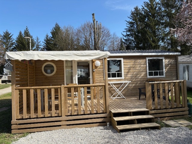 The image depicts a wooden mobile home set in a green outdoor environment. A covered terrace is visible at the front, featuring a seating area and a small table. Steps lead from the terrace to the gravel path, while windows allow natural light into the interior.
