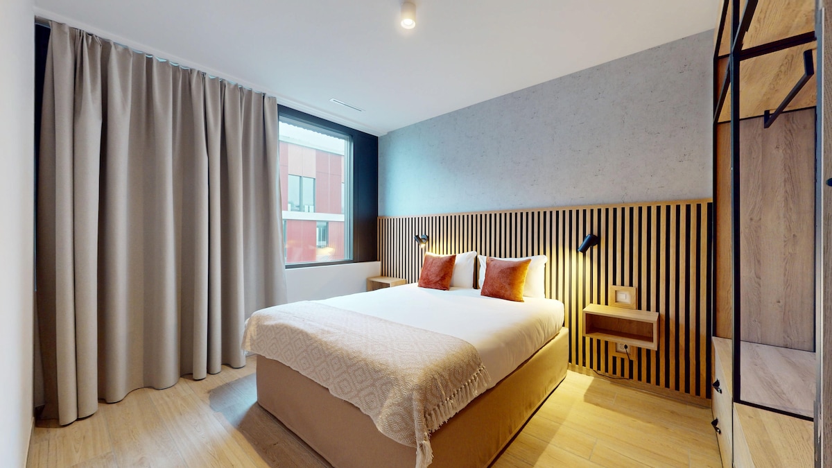 A well-lit bedroom showcases a spacious bed with a light-colored blanket, framed by wooden wall panels. Two decorative pillows in earthy tones sit atop the bed. A large window, covered by beige curtains, lets in natural light, highlighting the warm wooden flooring.