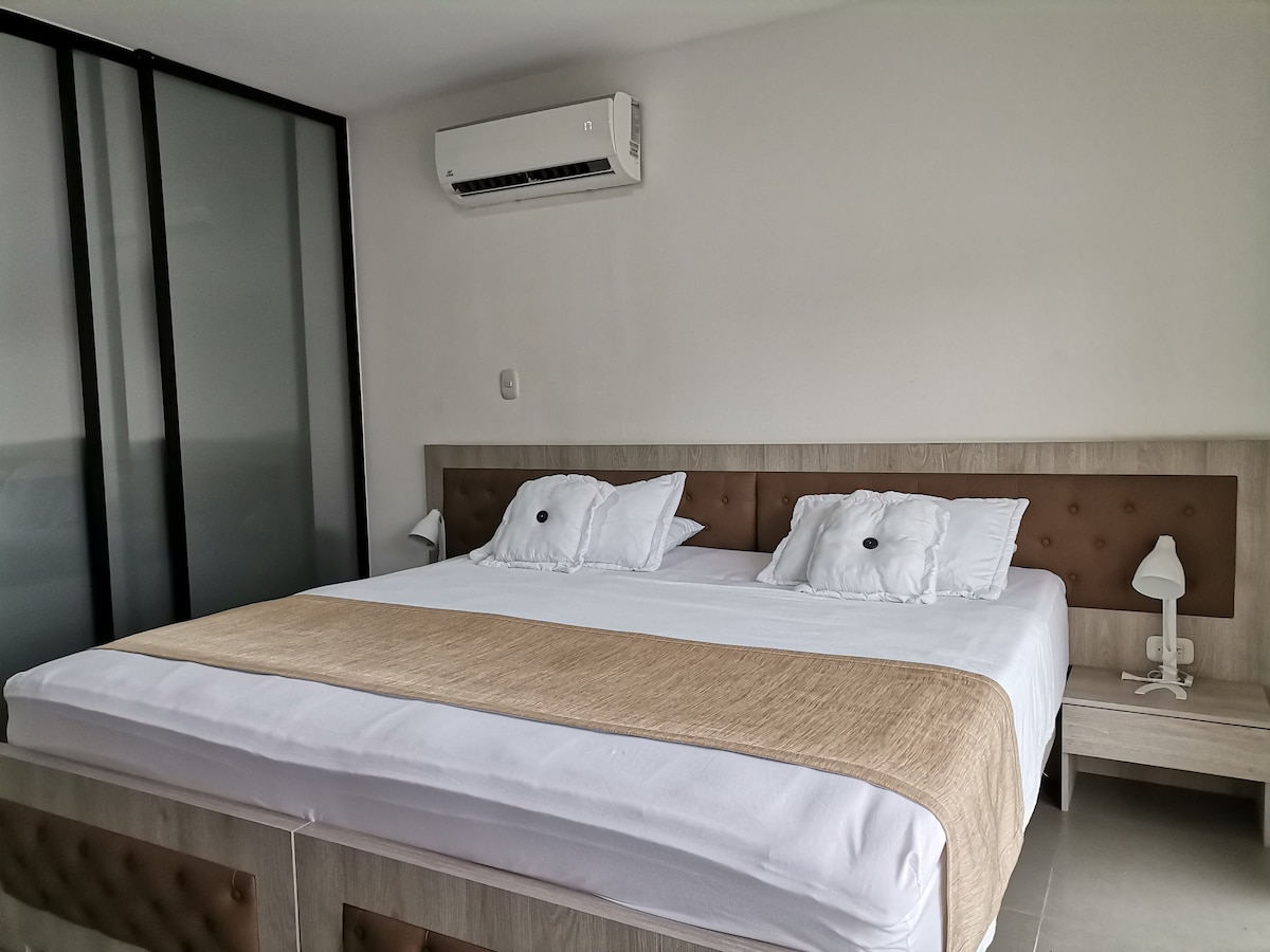 A spacious sleeping area features a king-size bed with decorative pillows. A bedside table and lamp are placed next to the bed. A modern air conditioning unit is mounted on the wall. The room is designed with natural light flowing in from a large window, enhancing the serene ambiance.