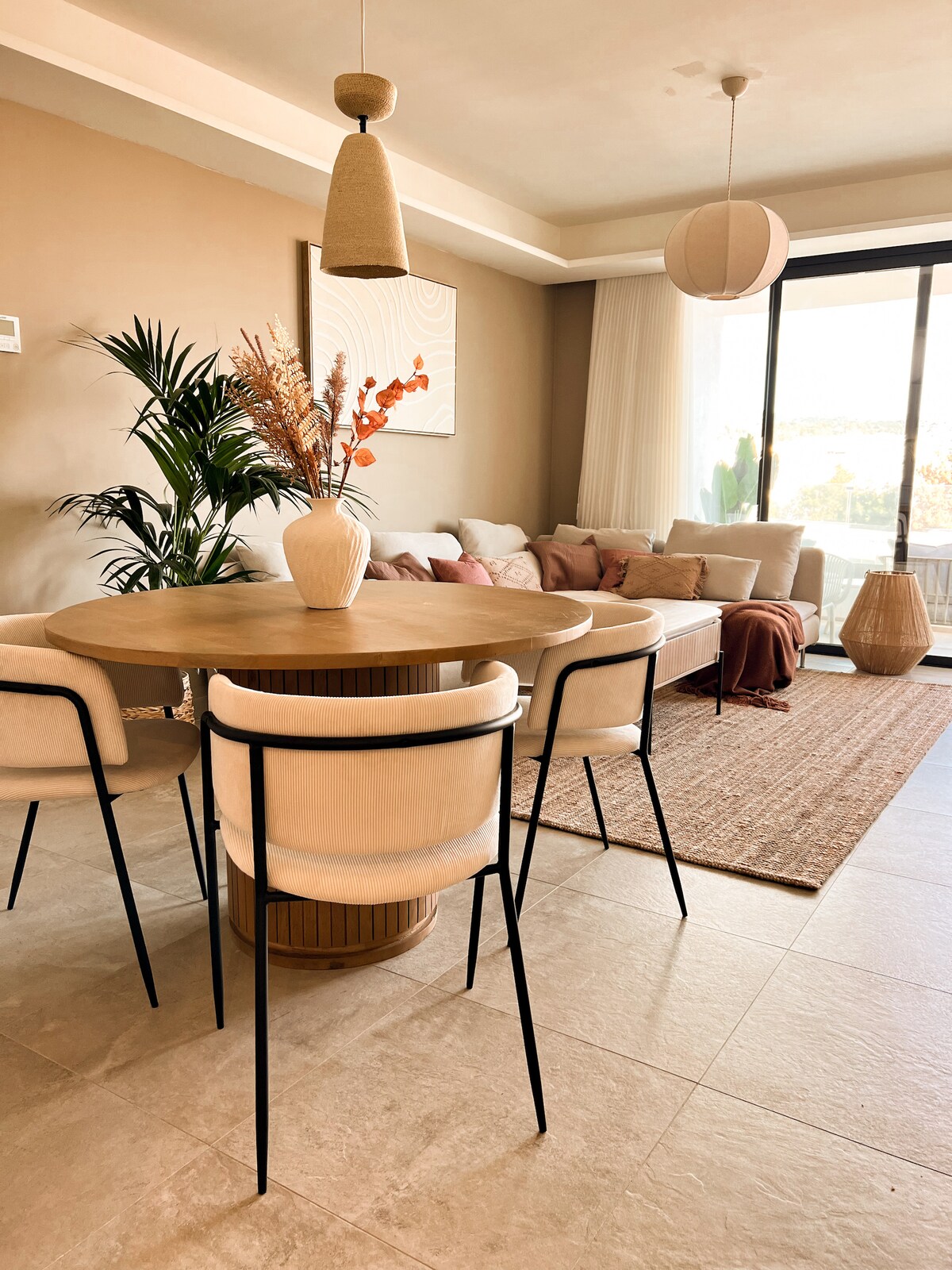 A dining area features a circular wooden table surrounded by four light-colored chairs. A decorative vase with dried flowers sits at the center. Soft furnishings in the background include a cozy sofa with various cushions, and large glass doors allow natural light to fill the space.