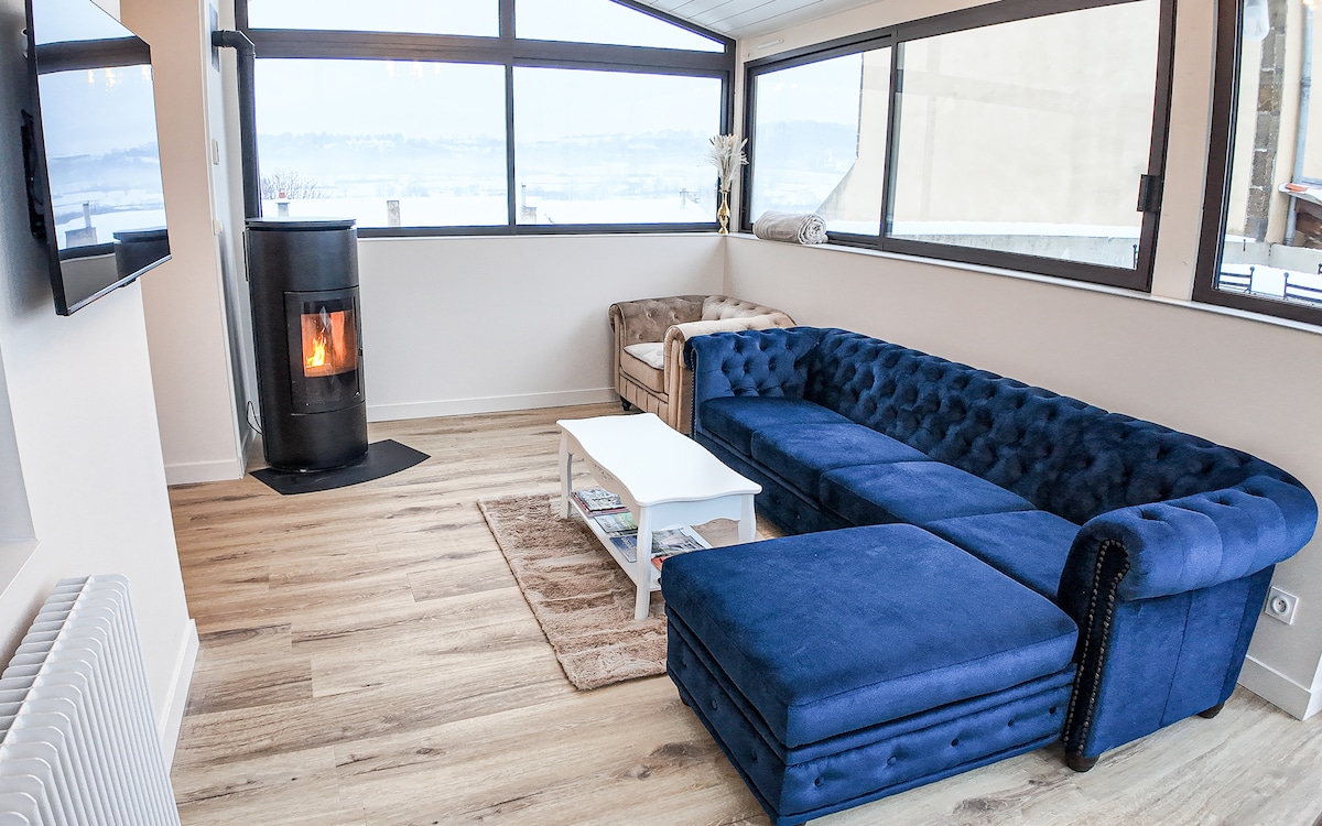 A light-filled living area is presented, featuring a deep blue sectional sofa and a plush armchair. A small coffee table sits in front of the sofa, while a sleek pellet stove provides warmth. Large windows offer scenic views and natural light.