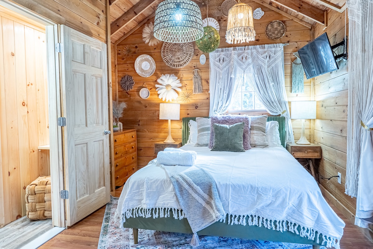 A cozy bedroom with wooden walls features a spacious bed adorned with neutral linens and decorative pillows. Two woven pendant lights hang overhead, and a television is mounted on the wall. A door leads to the bathroom, providing access to additional amenities.