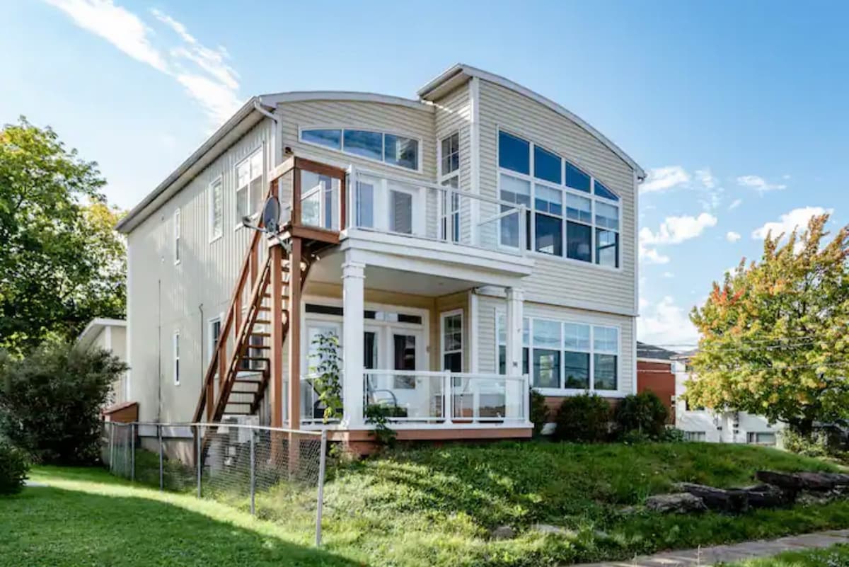 The exterior of a spacious two-story apartment is highlighted, featuring large windows that allow ample natural light. A welcoming front porch with a railing is visible, accompanied by a staircase leading to the main entrance. Lush green grass surrounds the property, complemented by nearby trees.