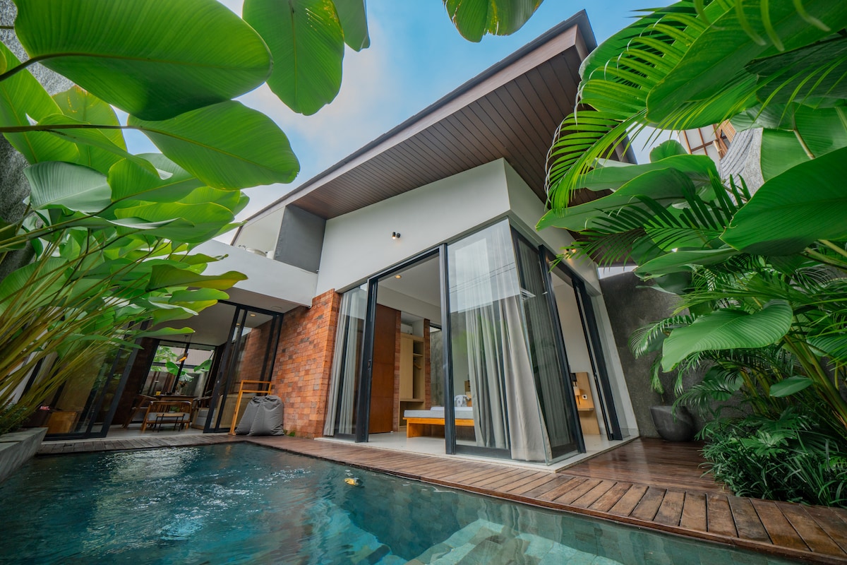 A modern villa is framed by lush tropical plants, featuring a private pool with wooden decking. Large glass doors open to an inviting interior, where natural light enhances the warm tones of the space. The serene ambiance is complemented by the surrounding greenery.
