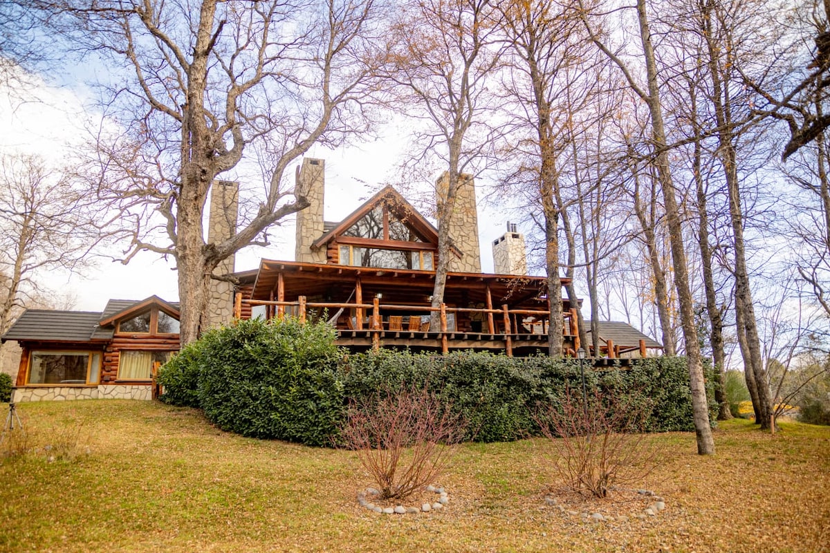 A charming wooden cabin is nestled among tall trees, showcasing a spacious deck that wraps around the structure. The natural landscape features a well-maintained lawn with shrubs, creating a serene setting. Stone chimneys rise from the roof, complementing the rustic design.