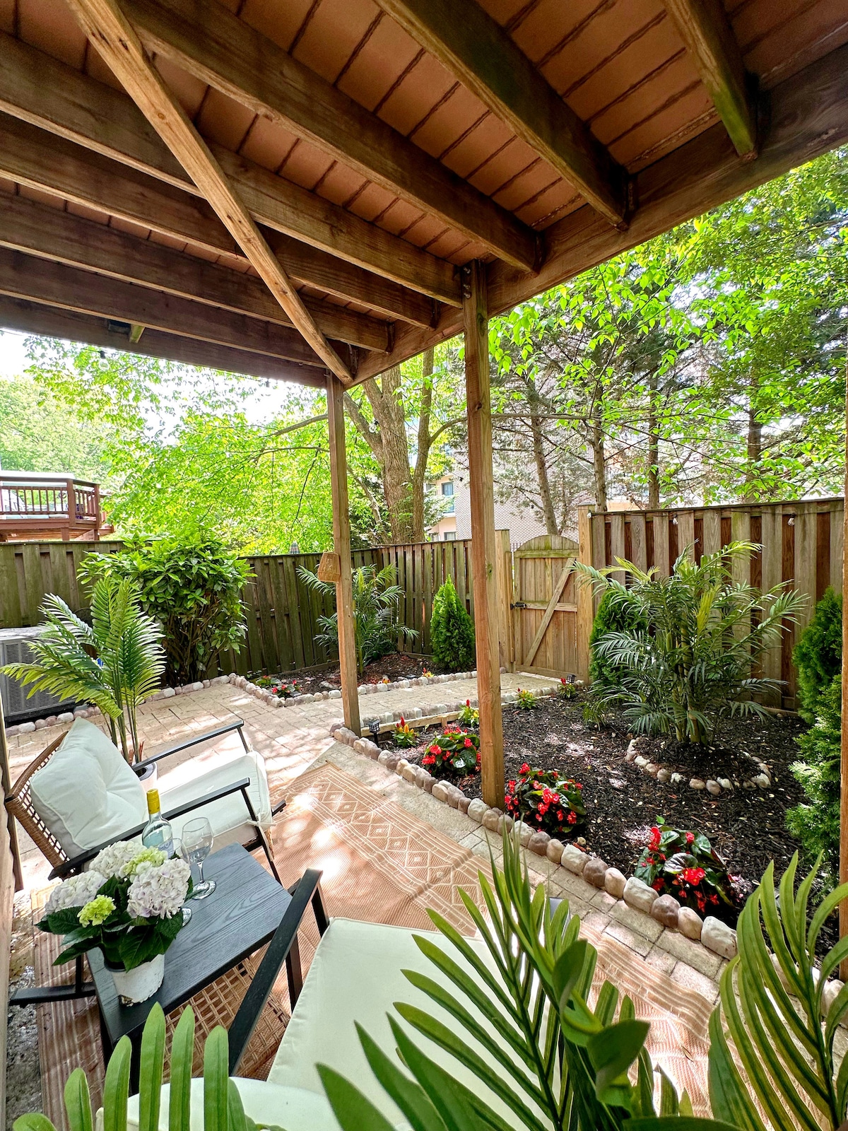 A private backyard oasis is displayed with shaded seating areas featuring light-colored chairs and a wooden table. Lush green plants and colorful flower beds enhance the natural surroundings, while a flagstone path leads towards a wooden gate, creating a serene outdoor space.