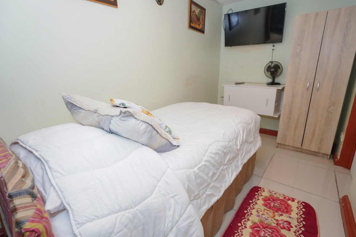 A cozy individual room features a spacious bed with a white comforter and a decorative pillow. A wall-mounted TV is positioned above a simple white cabinet. A wardrobe offers additional storage, and a patterned rug adds a touch of warmth to the tiled floor.