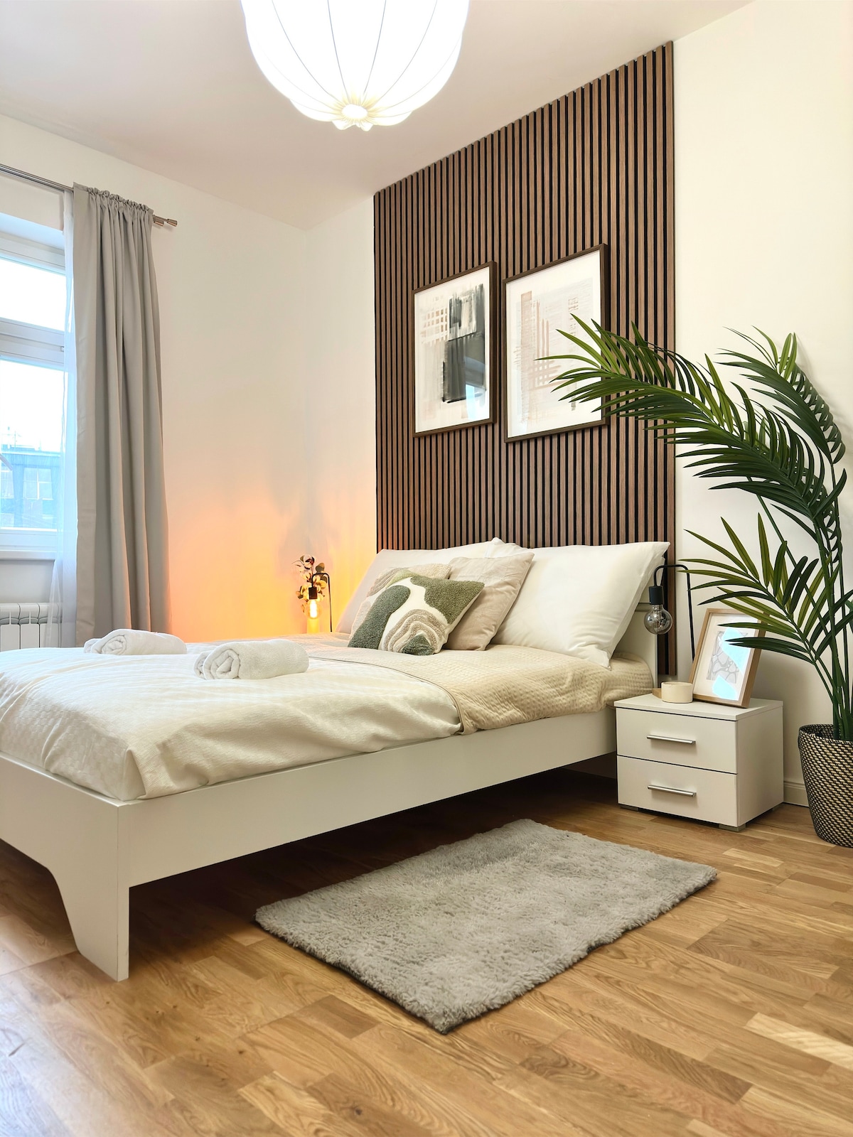 A cozy bedroom features a spacious bed dressed with neutral linens and decorative pillows. A small nightstand with a lamp sits beside the bed, while a potted plant adds a touch of greenery. Soft light illuminates the room, enhancing its relaxing ambiance.