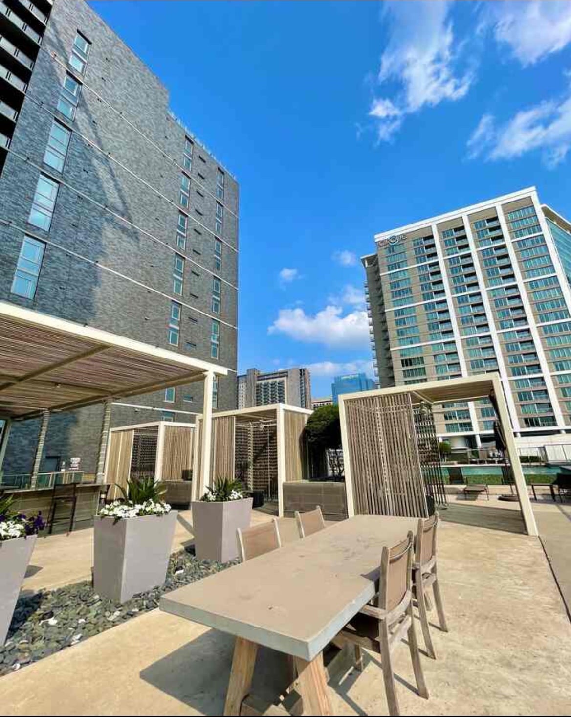 An outdoor dining area features a long wooden table surrounded by several chairs, with planters showcasing green plants and flowers. The backdrop includes modern high-rise buildings against a clear blue sky, creating a spacious and inviting atmosphere.