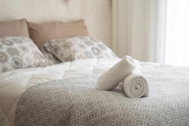 Habitación elegante con un ambiente suave y cálido, con ropa de cama mullida con una colcha con motivos sutiles, almohadas a juego y toallas blancas cuidadosamente dobladas para una presentación acogedora.