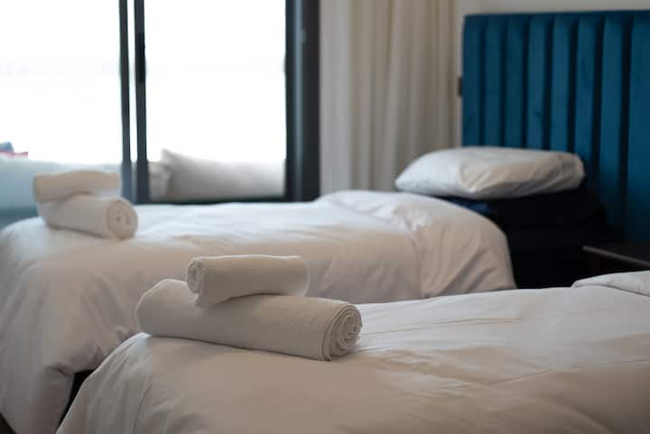 Habitación luminosa con dos cómodas camas individuales,ropa de cama blanca brillante, elegante cabecero de terciopelo azul y gran ventanal con vistas naturales. Un espacio refinado y sereno, ideal para descansar y disfrutar del entorno circundante