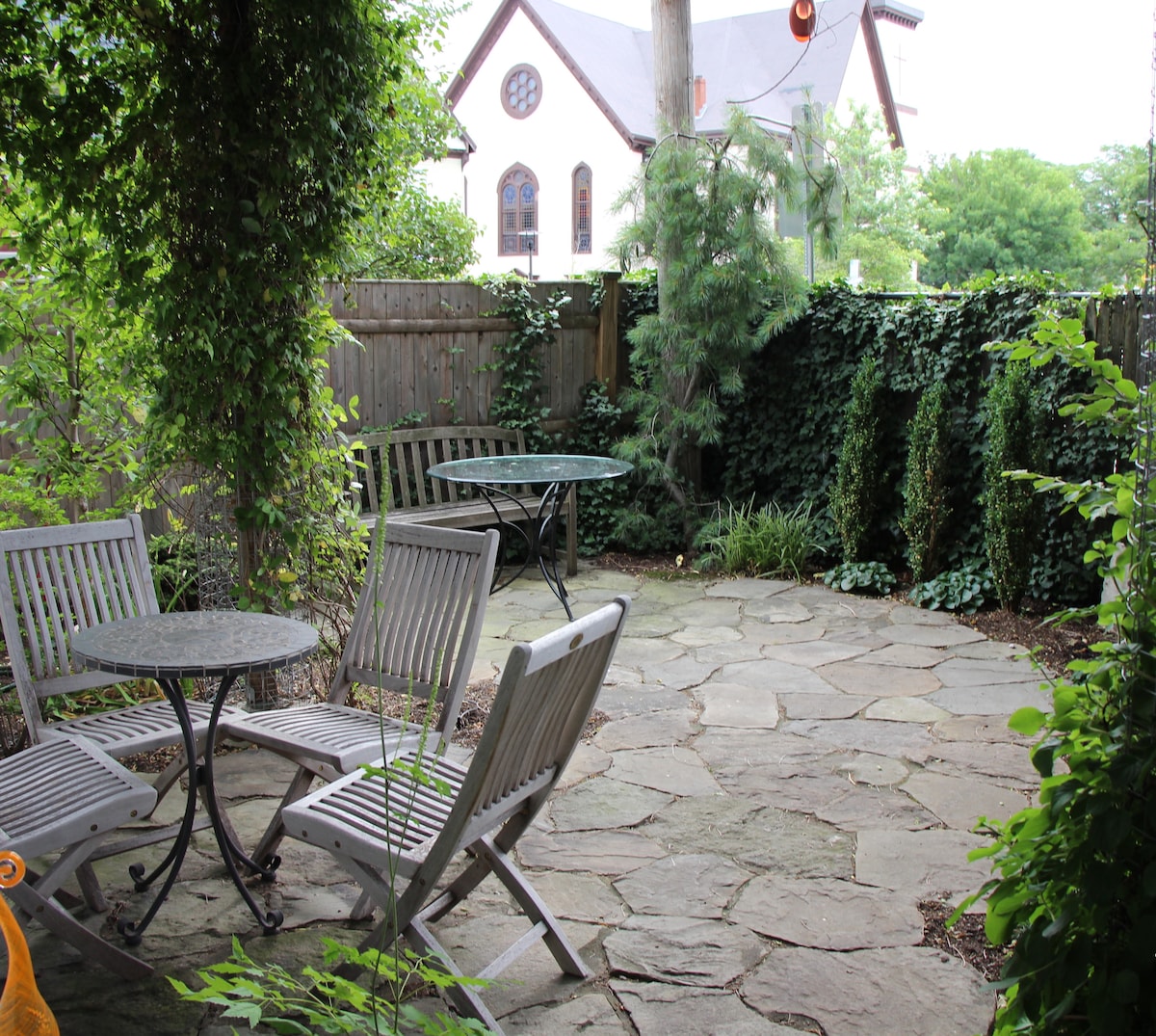 A peaceful garden oasis is presented, featuring stone paving and surrounded by lush greenery. Two round tables are set among wooden chairs, inviting moments of relaxation. A backdrop of decorative shrubs and climbing plants enhances the tranquil ambiance of the outdoor space.