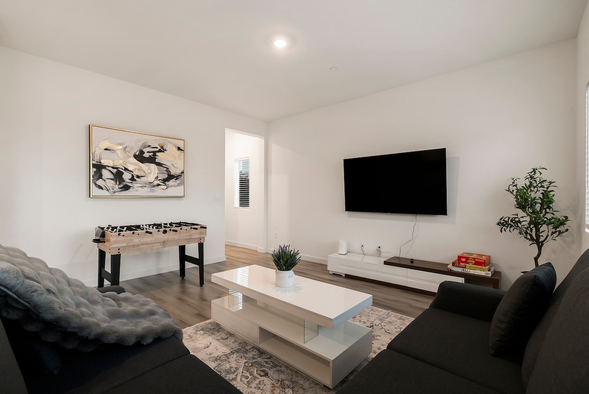 A modern living area features a sleek black sofa and a coffee table, complemented by a minimalist entertainment unit and a decorative area rug. A game table is positioned to the side, while natural light is provided by a nearby window.