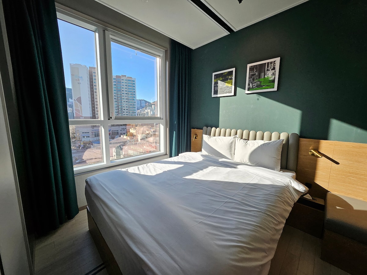 A bright and cozy room features a spacious bed with crisp white bedding. Large windows allow natural light to illuminate the space, showcasing green accent walls and framed photos. A bedside table with a lamp completes the inviting atmosphere.