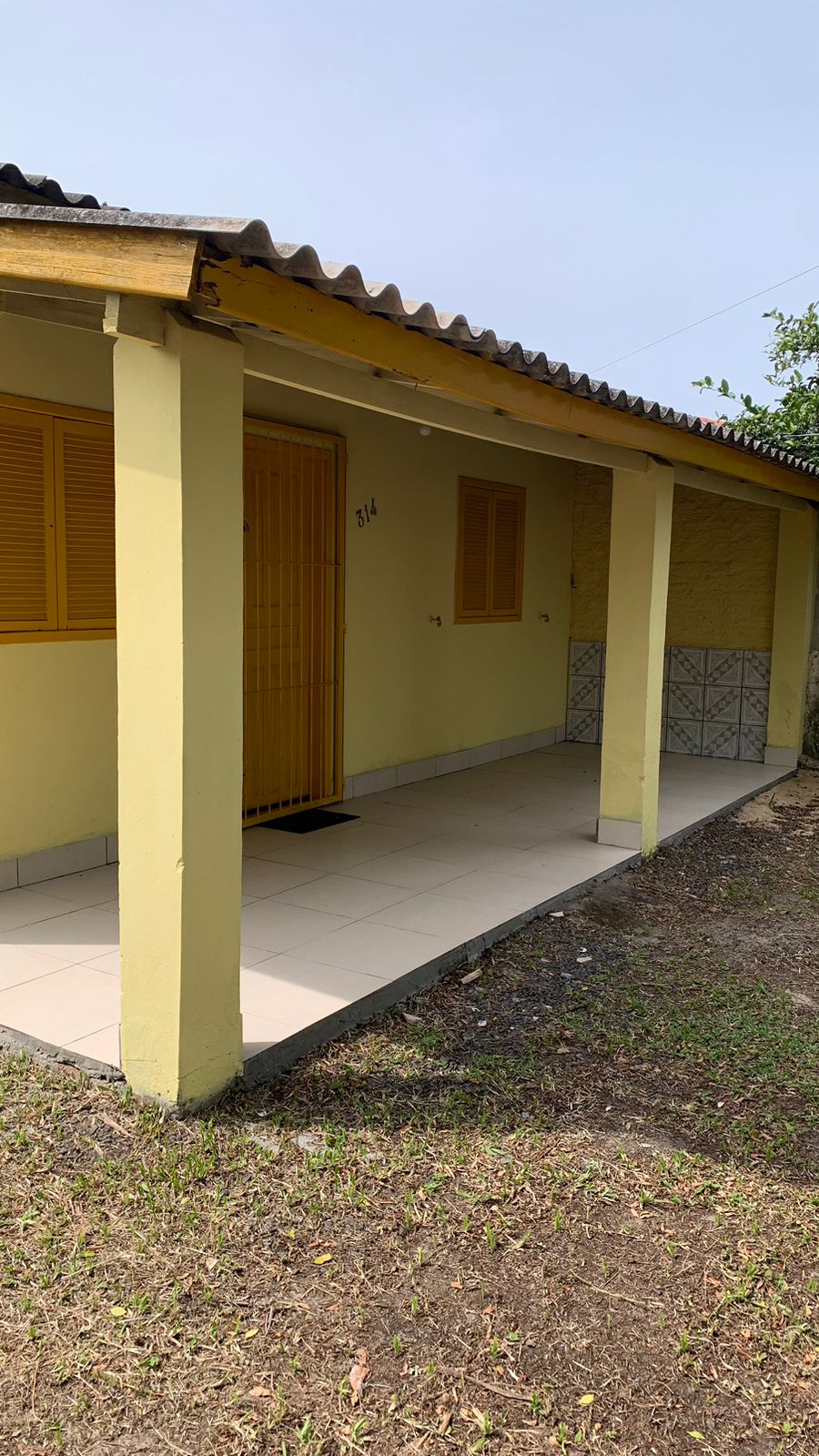 A small yellow cottage with a welcoming entrance is depicted. The porch features tiled flooring and is complemented by two windows with wooden shutters. The surrounding area consists of trimmed grass, suggesting a relaxed outdoor environment.