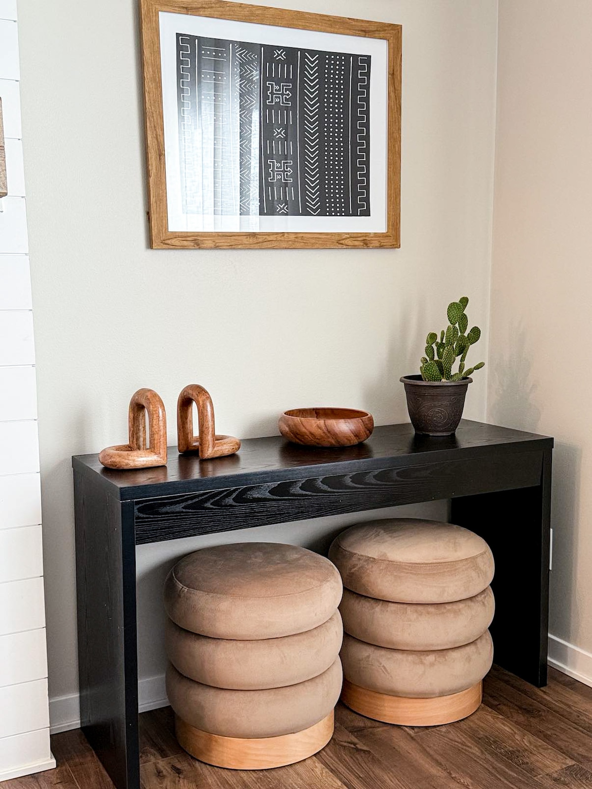 A narrow black console table is positioned against a light wall. Above it, an artwork in a wooden frame features geometric patterns in black and white. Two wooden sculptures and a wooden bowl are placed on the table, accompanied by a small potted cactus.
