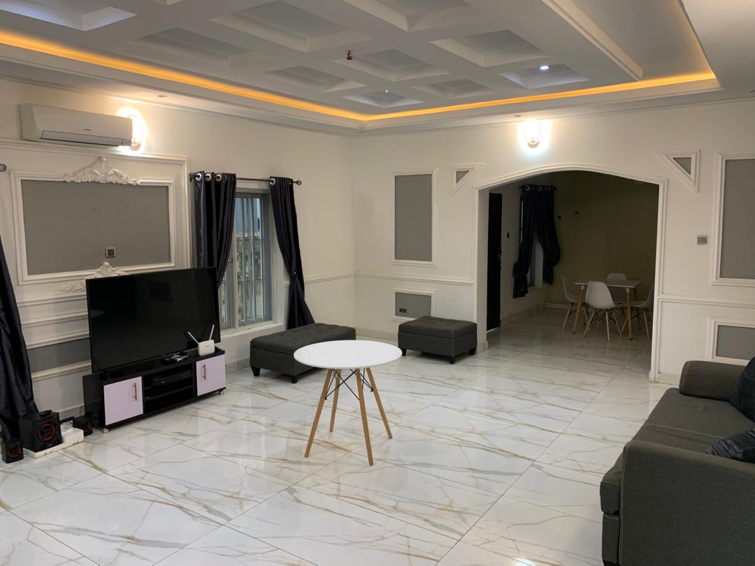 A spacious living room is presented with modern furnishings, including a dark sofa and a white table supported by wooden legs. A flat-screen TV is positioned against a light grey wall, while large windows with dark curtains allow natural light to enter the room.