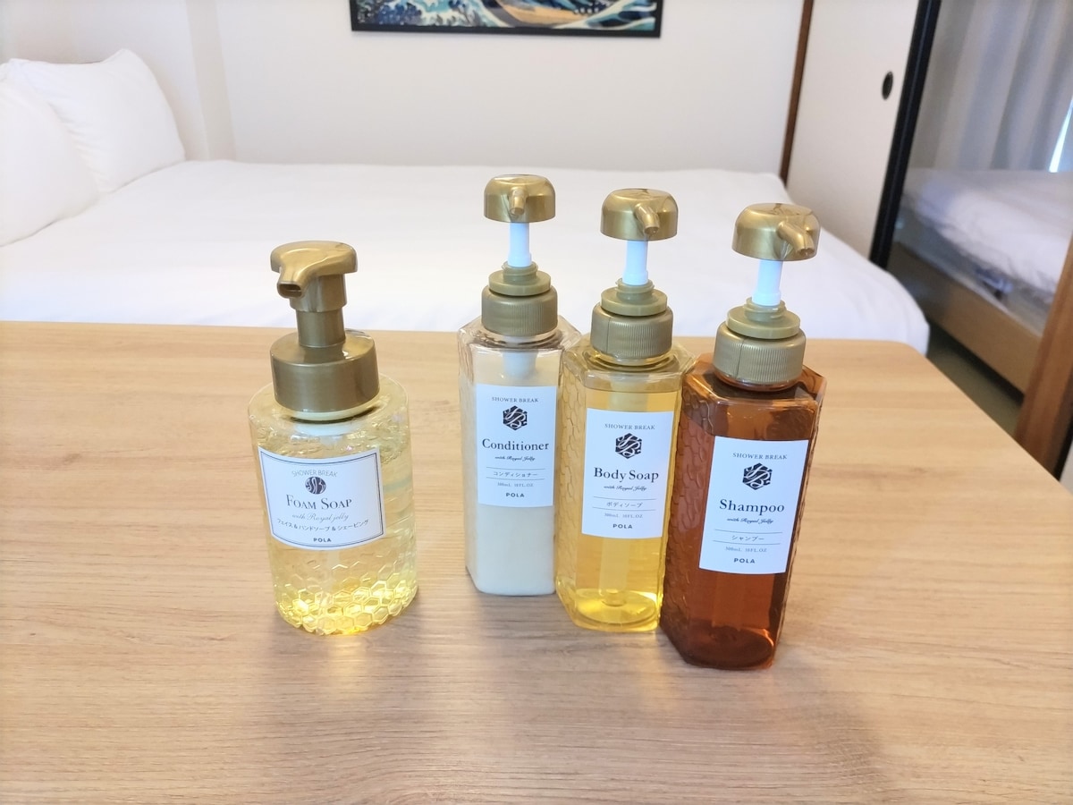 Four pump dispenser bottles are neatly arranged on a wooden surface. The bottles contain foam soap, conditioner, body soap, and shampoo, all branded with the POLA label. A spacious bed can be seen in the background, contributing to a relaxed atmosphere.