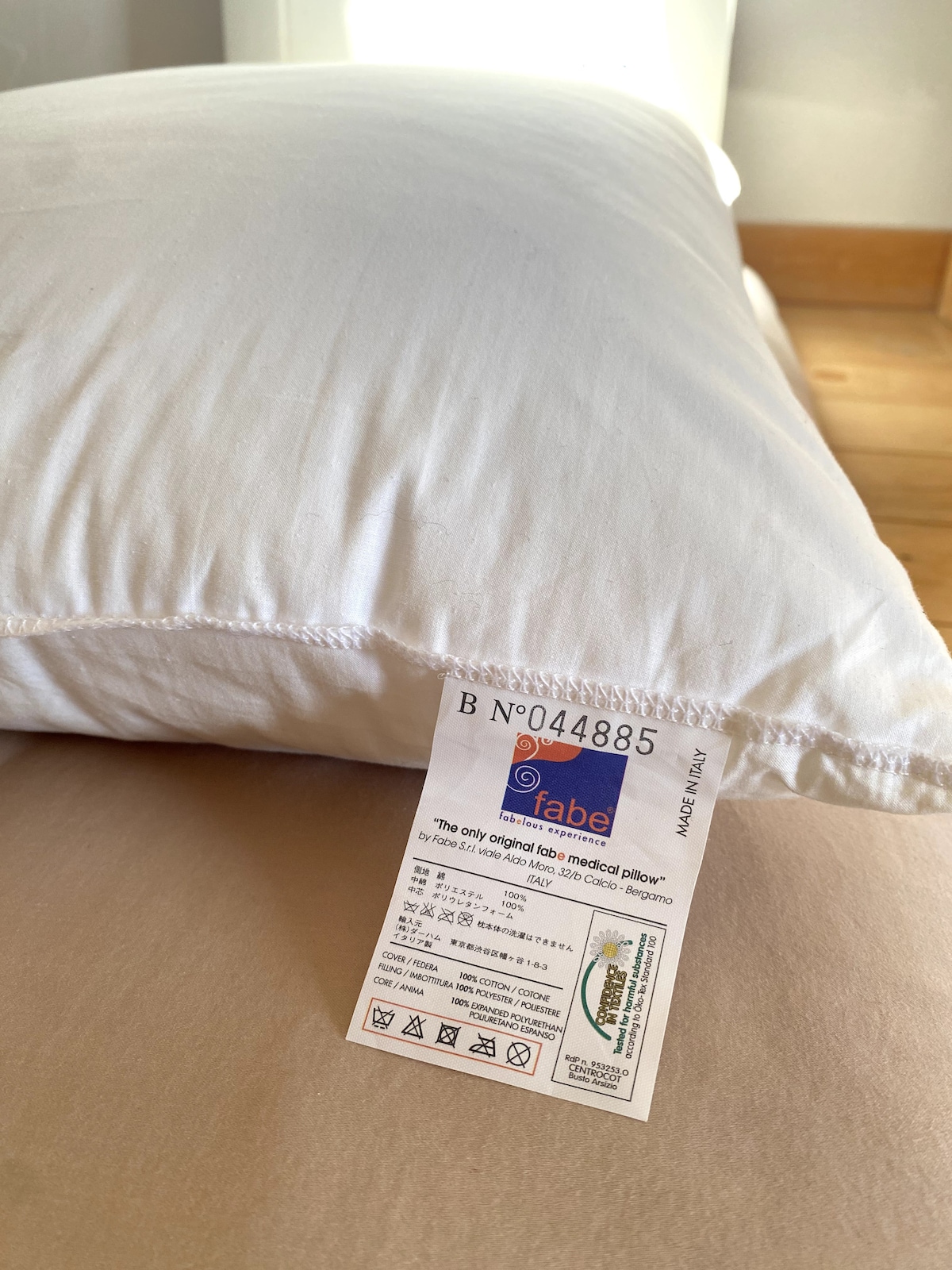 A soft, white pillow is displayed on a comfortable bed. A care label is visible, providing information about the pillow's material and maintenance. The neutral-colored bedding complements the cozy setting of the room.