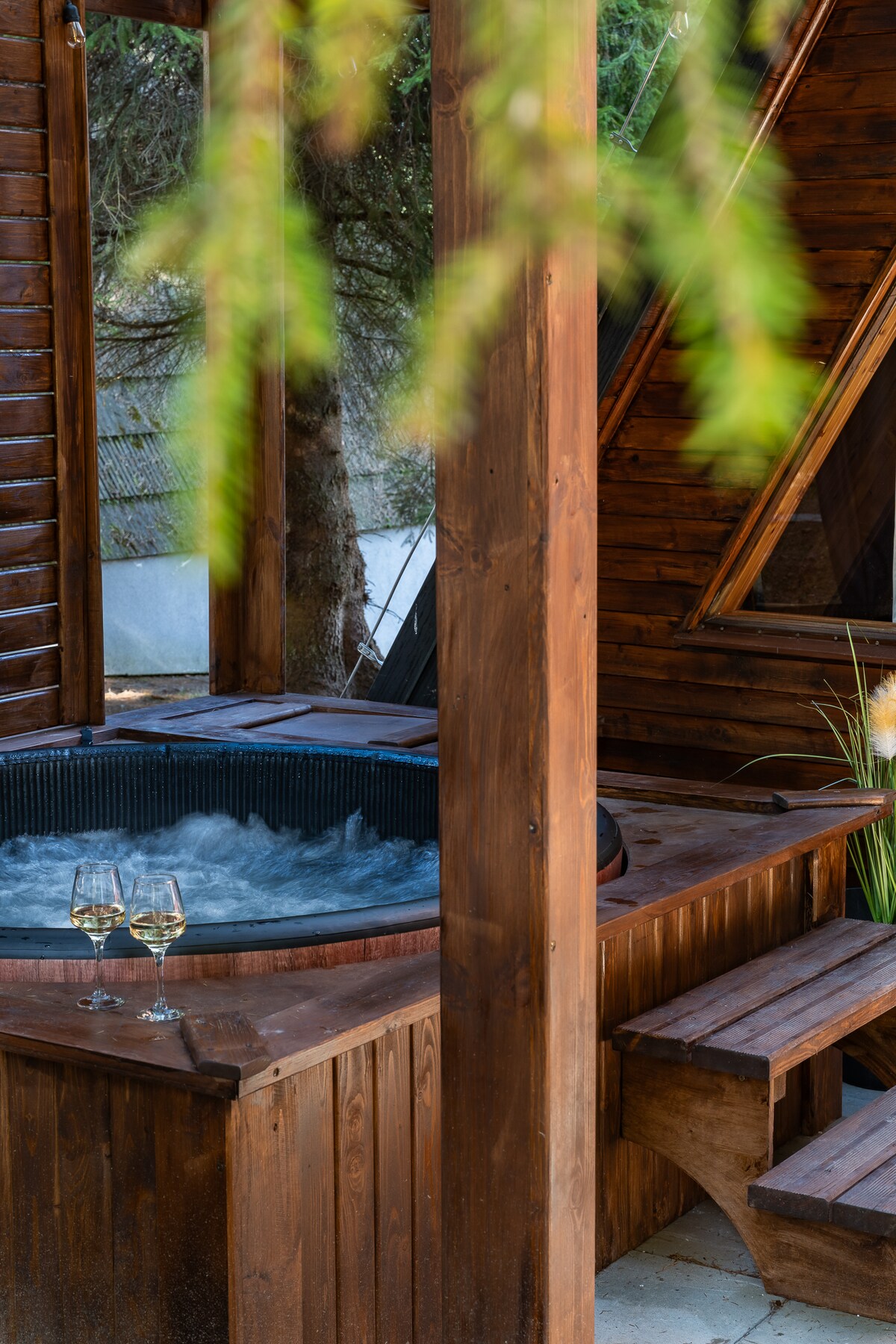 A private jacuzzi is situated on a wooden deck, surrounded by lush greenery. Two glasses are placed nearby, suggesting a tranquil setting. The structure features wooden walls and an angled roof, creating an inviting atmosphere.