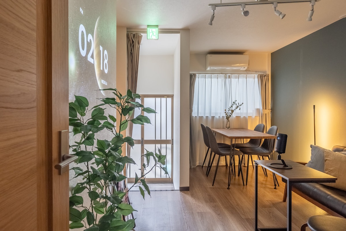 A well-lit living area features a dining table with four chairs and a cozy sofa. A projection displaying the time occupies one wall. Light streams through a large window with sheer curtains, creating a welcoming ambiance. A potted plant adds a touch of greenery.