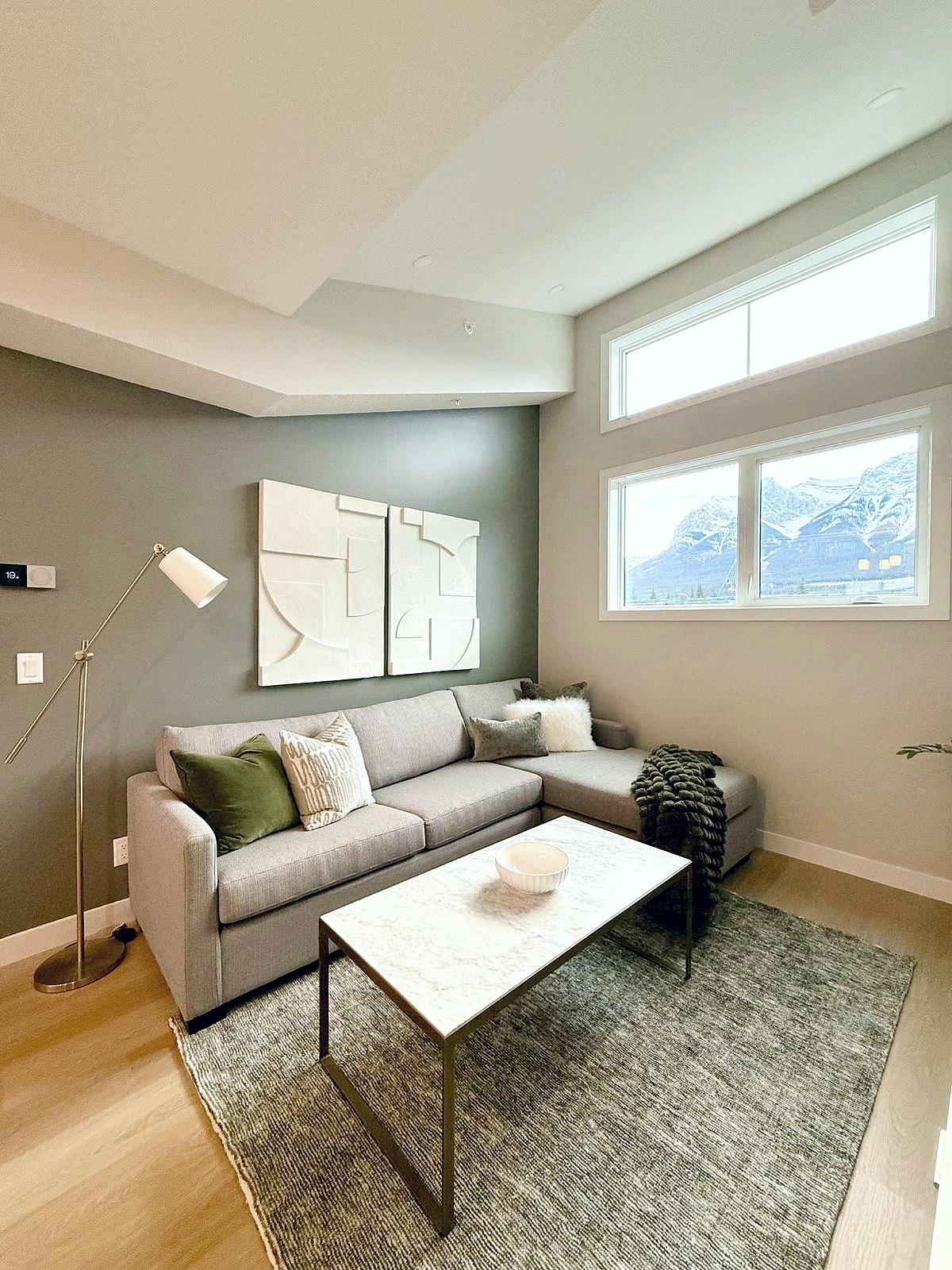 A cozy living area is showcased, featuring a light gray sofa adorned with textured cushions. A coffee table rests on a soft area rug, with mountain views visible through large windows that provide natural light.