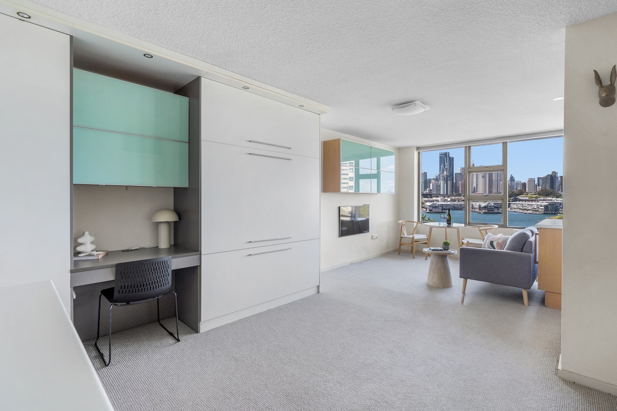 A bright studio space features a modern desk area with a stylish chair, complemented by a built-in cupboard. Large windows provide a panoramic view of Sydney's skyline and harbour, while light carpets and simple furnishings create a clean, contemporary aesthetic.